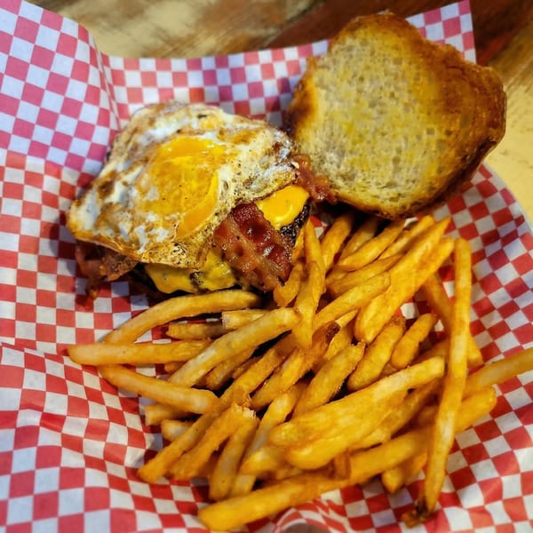 A bacon cheeseburger with a fried egg and a side of golden french fries in a red and white checkered basket.