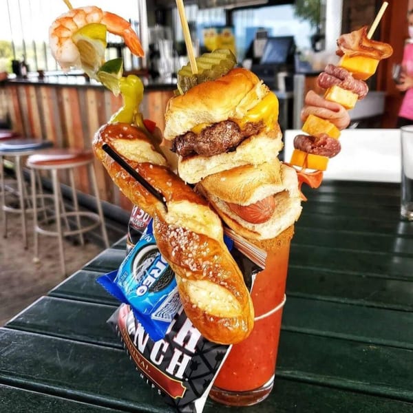 A Bloody Mary cocktail garnished with sliders, hot dogs, pretzel, shrimp, bacon, cheese, and chips.