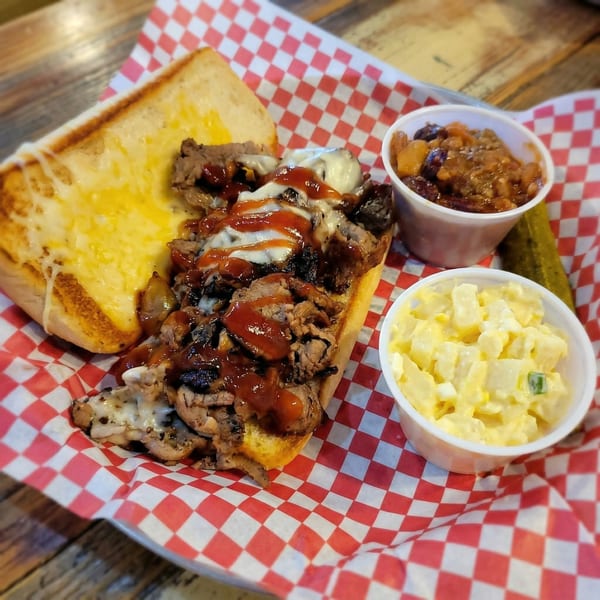 Cheesesteak sandwich with BBQ sauce, baked beans, potato salad, and a pickle on checkered paper.