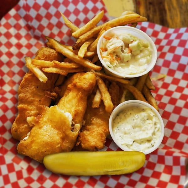 Golden fried fish and french fries with coleslaw, tartar sauce, and a pickle on red checkered paper.