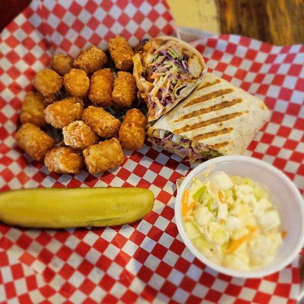 Grilled wrap with tater tots, coleslaw, and a pickle spear on red and white checkered paper.