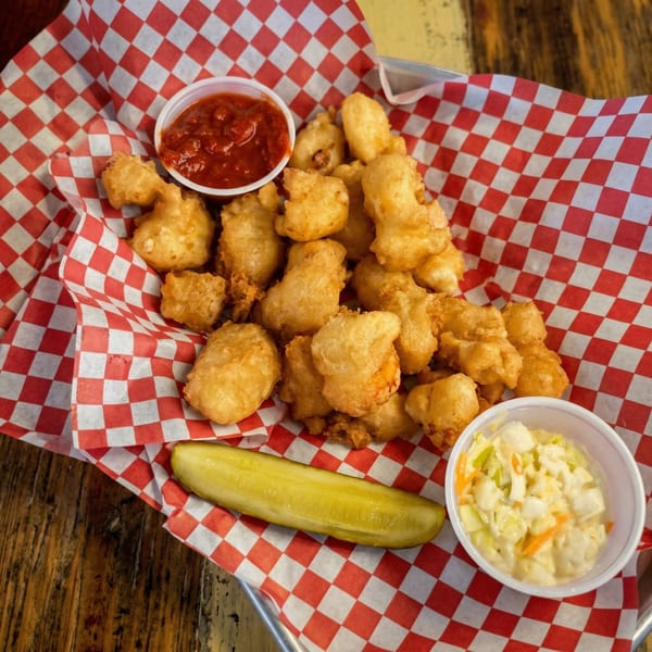 Golden-brown fried cheese curds with marinara sauce, coleslaw, and a pickle spear on checkered paper.