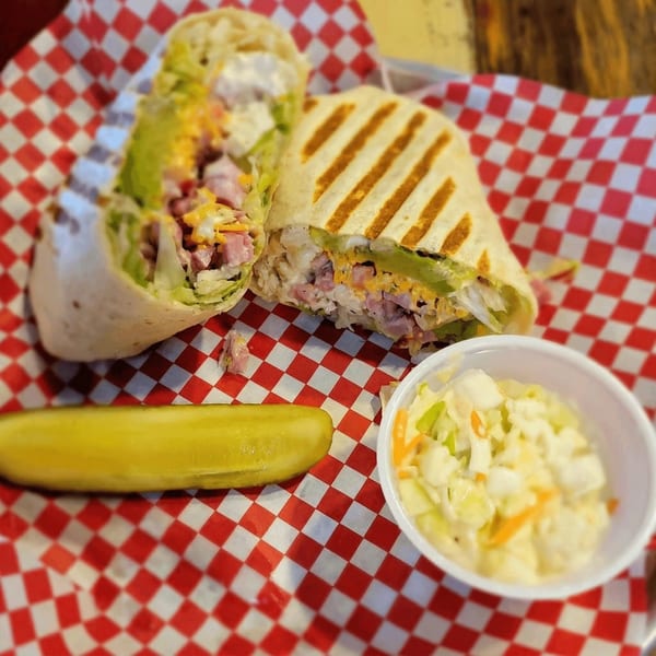 A grilled wrap, cut in half, revealing ham, cheese, and lettuce, served with a pickle and coleslaw.
