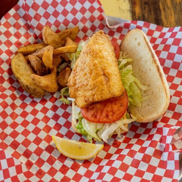 Fried fish sandwich with lettuce, tomato, and a side of potato wedges on red and white checkered paper.