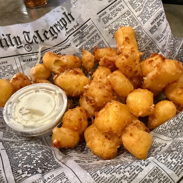 Golden-brown fried cheese curds and a side of dipping sauce served on newspaper-print paper.