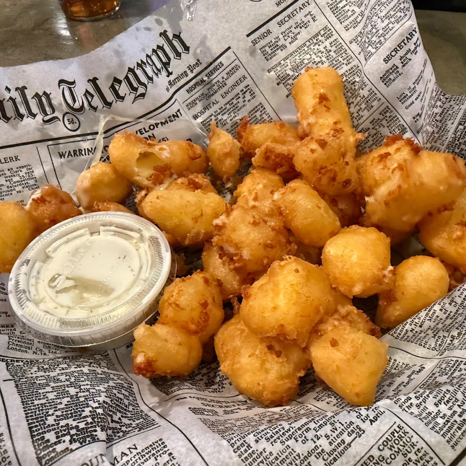 Golden-brown fried cheese curds and a side of dipping sauce served on newspaper-print paper.