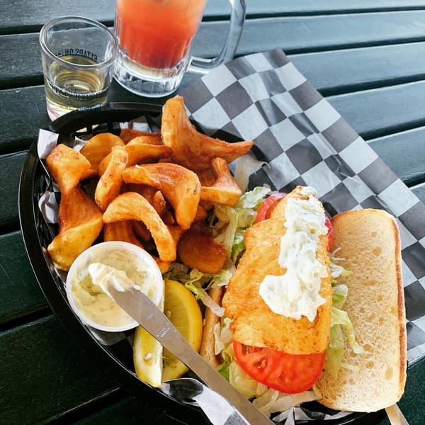 Fish sandwich with tartar sauce, lettuce, tomato, and a side of fries, lemon, and drinks.